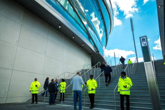 London, UK - 12 May, 2019: color image depicting teams of security workers wearing high-visibilty jackets working outside the brand new modern architecture of the Tottenham Hotspur stadium in north London, UK. It is the day of an English Premier League match (Tottenham v Everton) and the security staff are waiting to inspect and frisk many thousands of football supporters. Room for copy space.